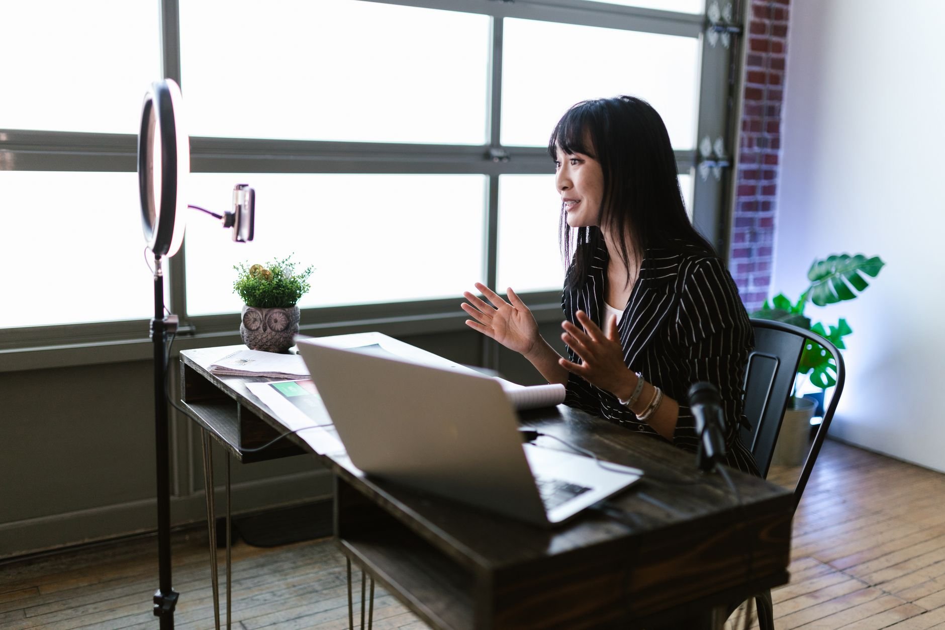 Woman gesturing while speaking to a laptop beside a ring light and microphone