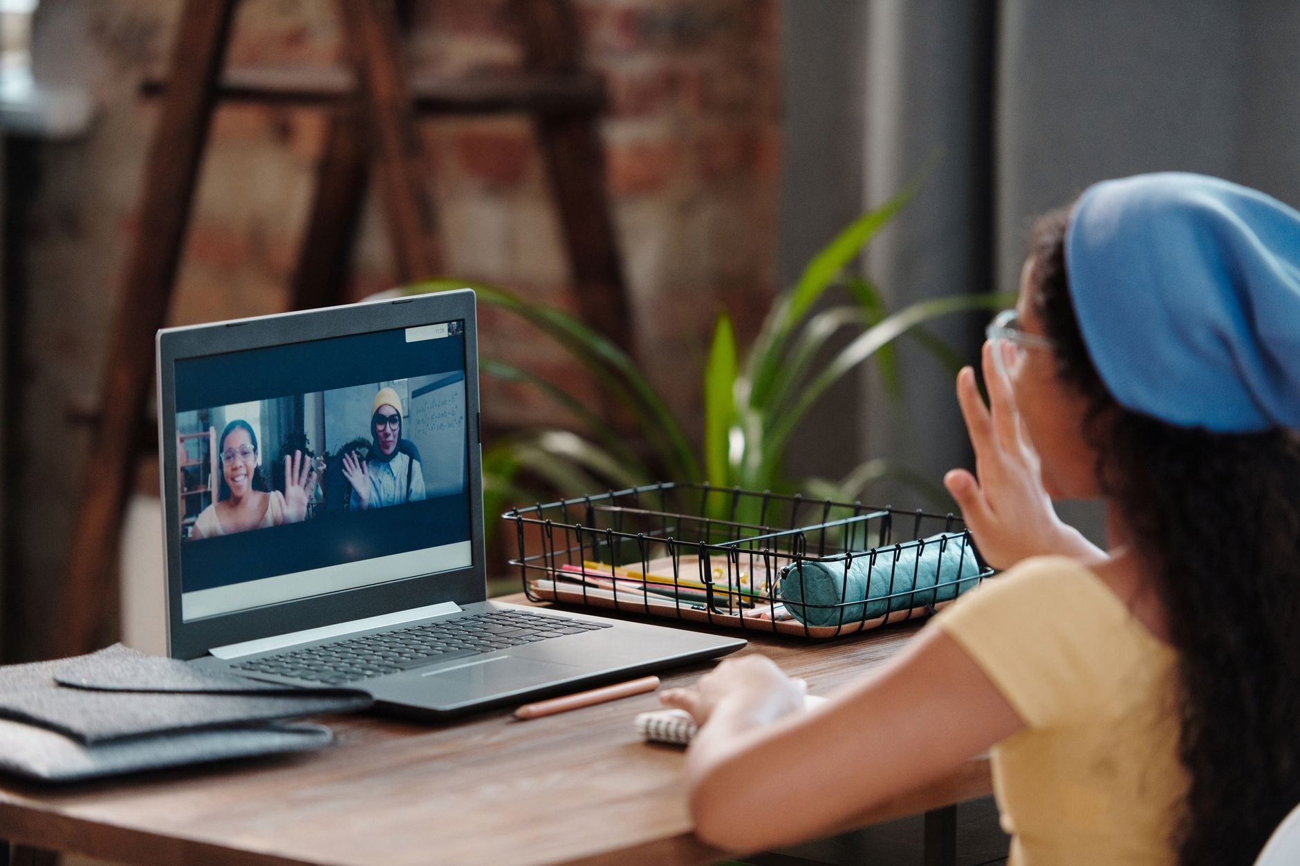 Woman in a yellow top and blue headscarf waving to people on a laptop video call