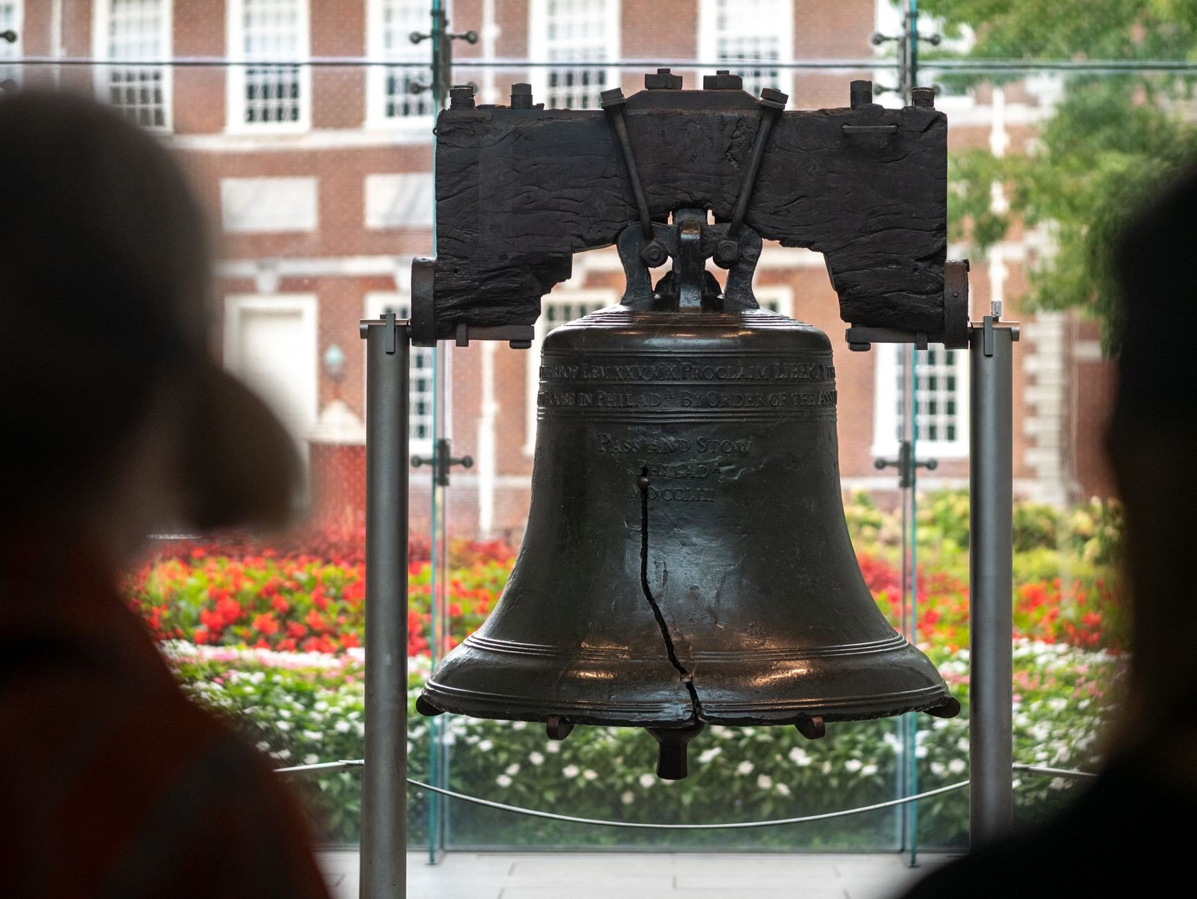 Liberty Bell with a visible crack, displayed in front of a brick building with red flowers, with two visitors in the foreground