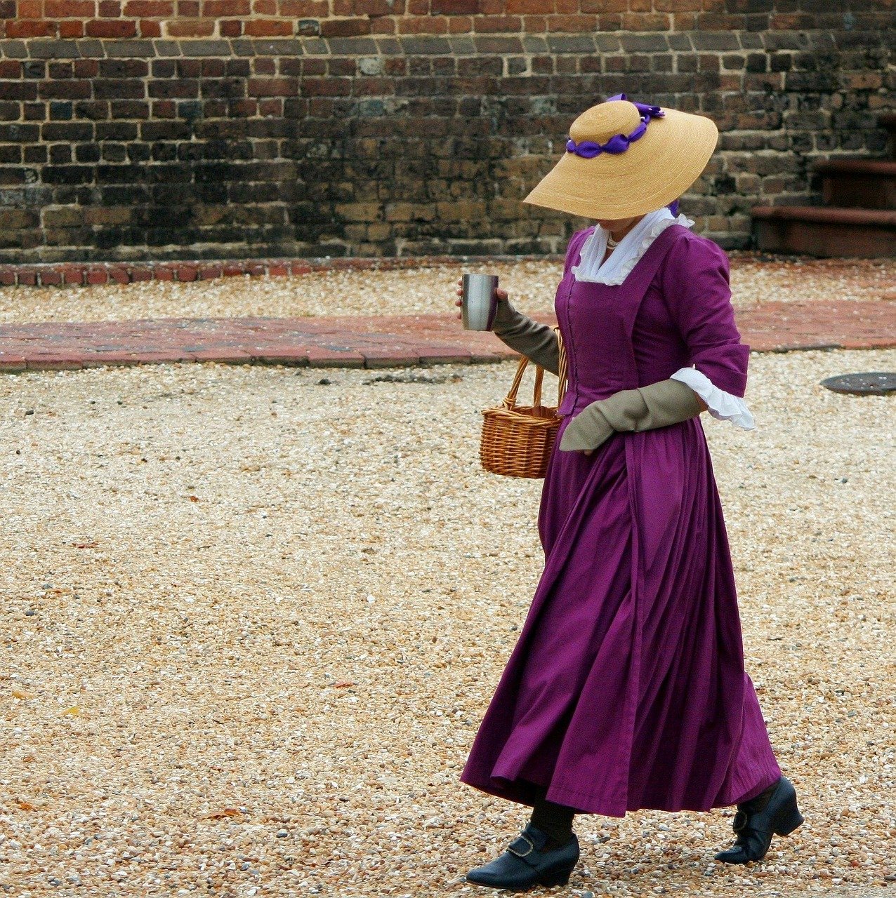 Woman in a purple 18th-century-style gown, straw hat with a purple band, and gray gloves, carrying a wicker basket and a mug on a gravel path beside a brick wall