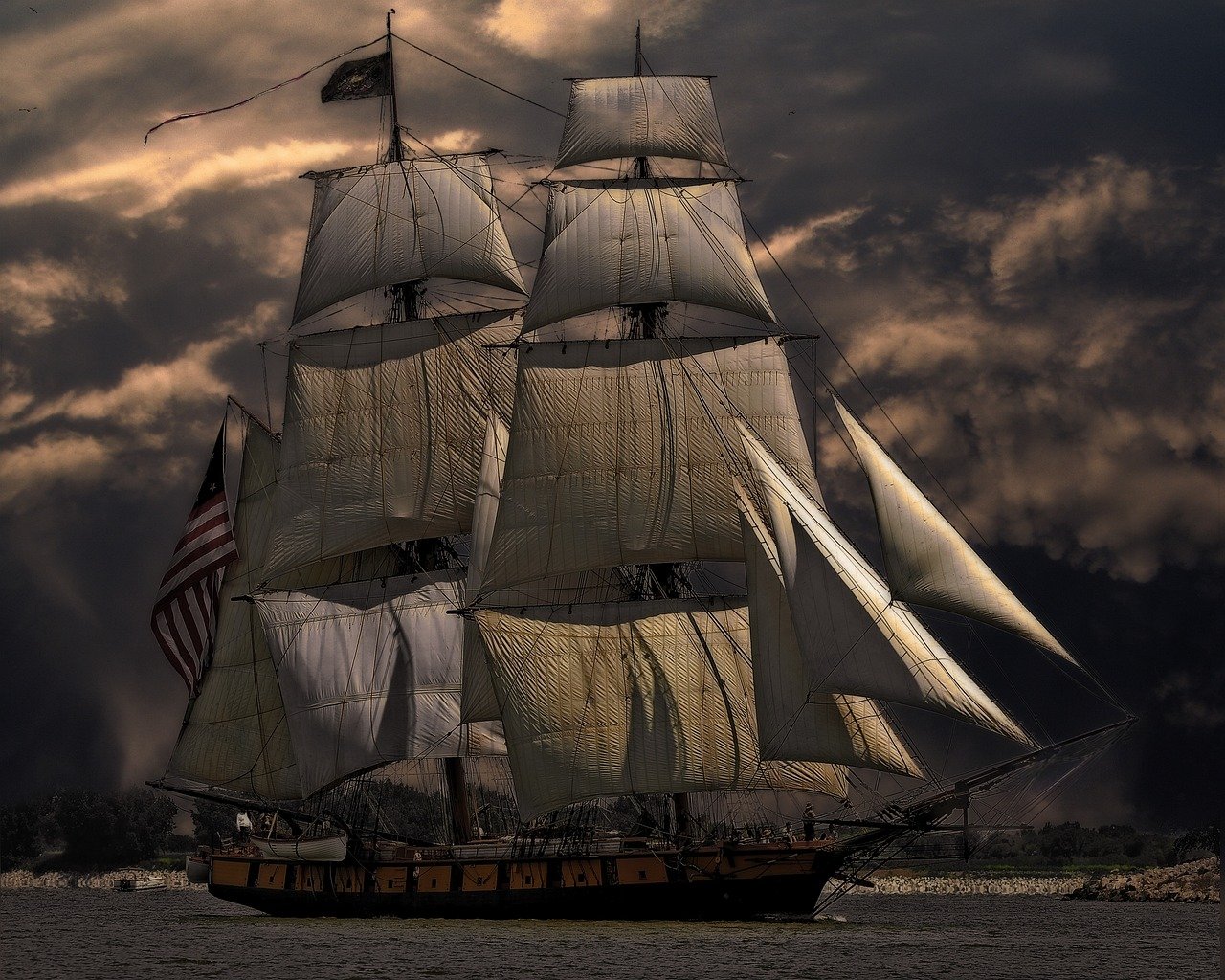 Large sailing ship with multiple tall sails and an American flag on the stern, under a dramatic cloudy sky above calm water