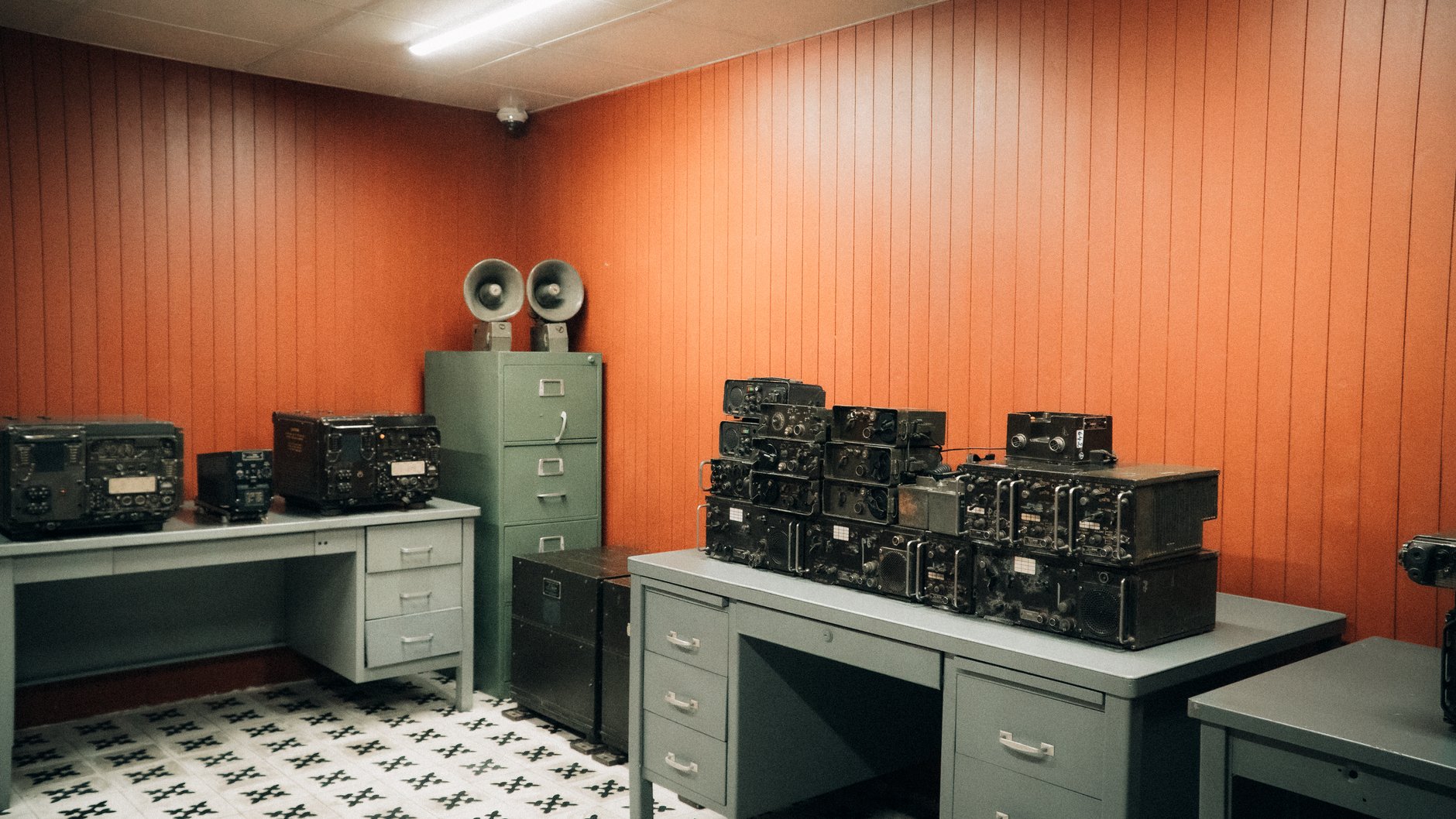 Room with orange wood-paneled walls holding several black vintage radio transceivers on gray metal desks and a filing cabinet topped with two horn speakers
