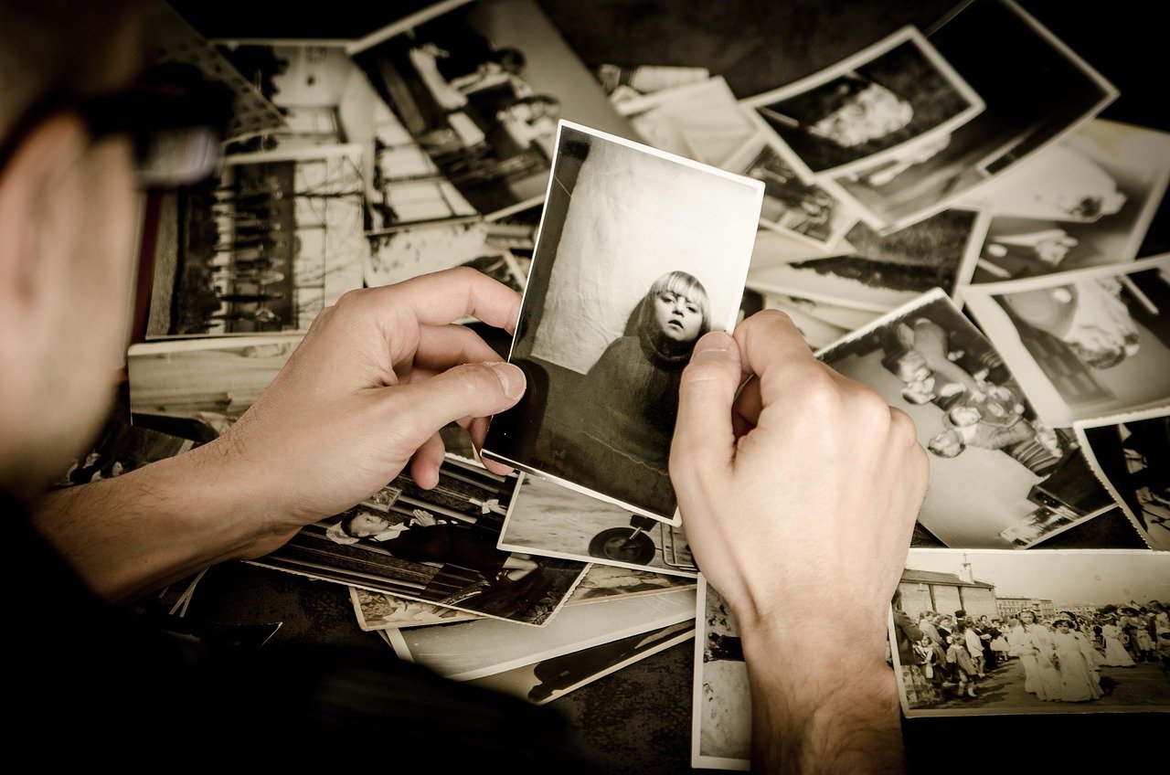 Hands sorting black-and-white vintage photographs spread across a table, with one print held up close to the camera