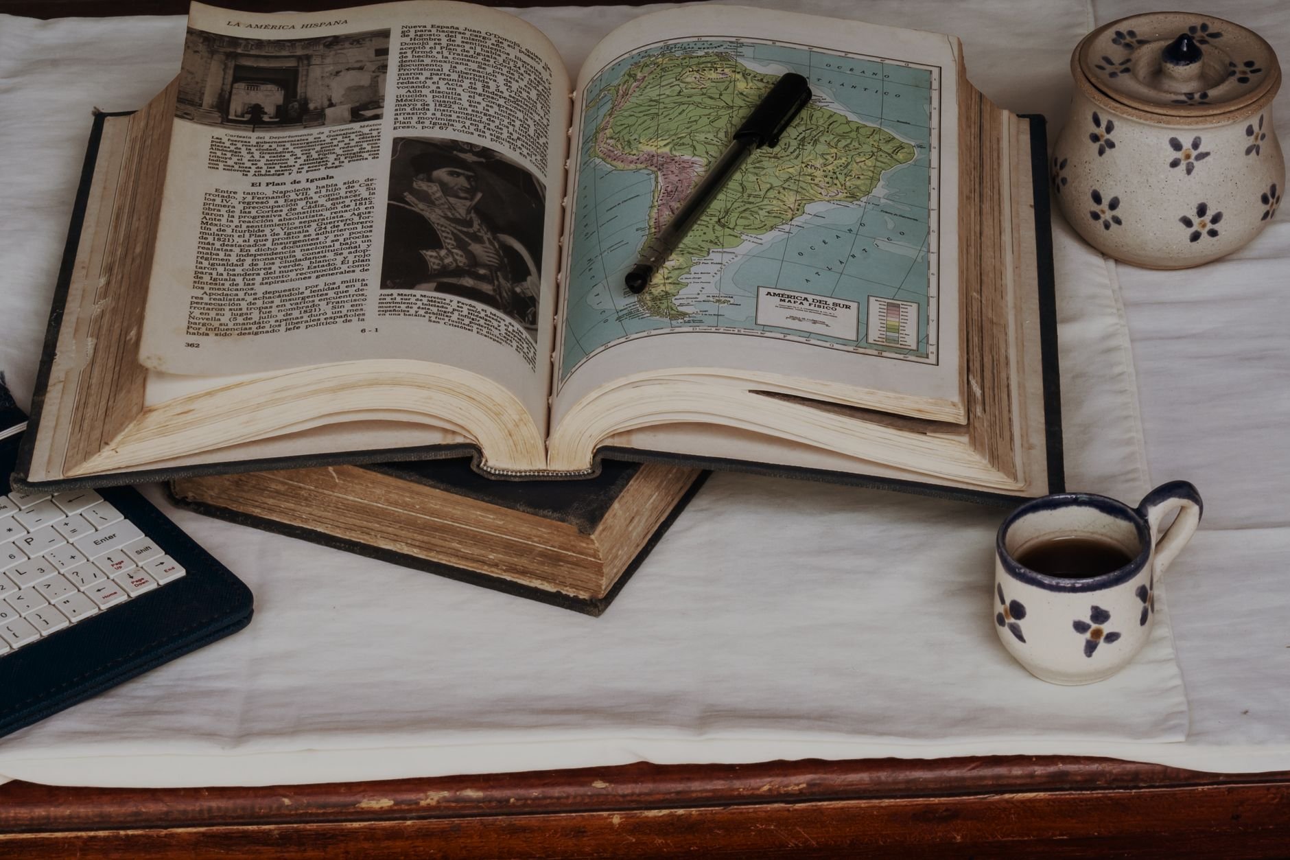 Open vintage book with a colored map of South America, a black pen resting on its pages, beside a small patterned cup and a patterned jar