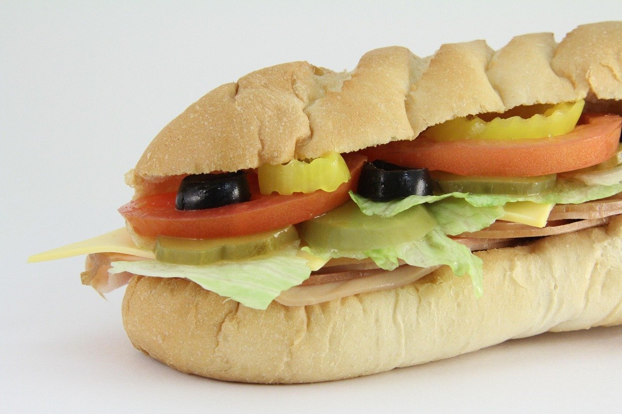 Long sub roll filled with sliced ham, cheese, lettuce, tomato slices, pickle slices, and black olives, on a white background