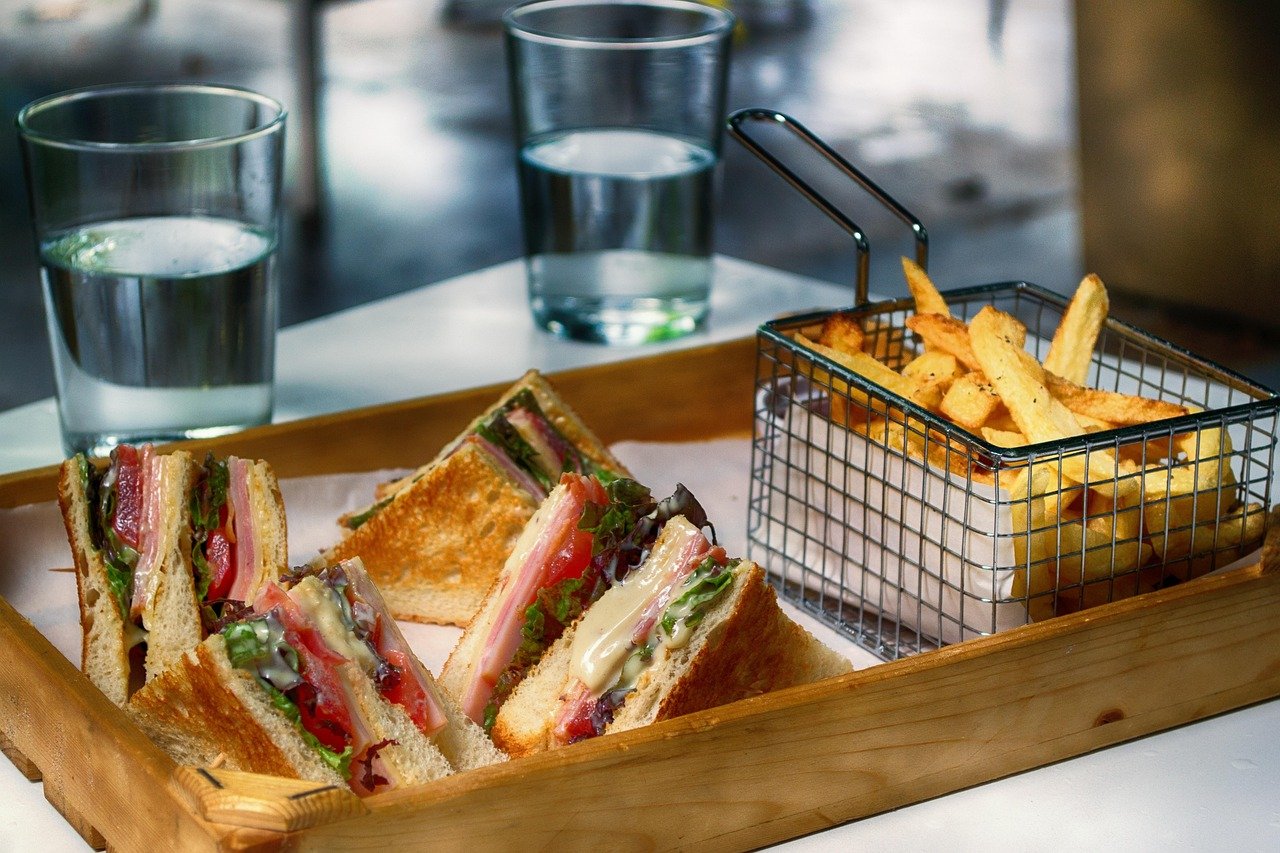 Triangle-cut club-style sandwich halves with layered meat, tomato, and greens on toasted bread, served on a wooden tray beside a wire basket of fries and two glasses of water