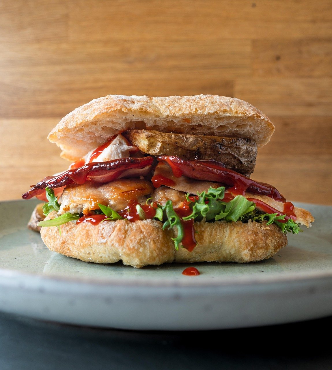 Thick sandwich on a toasted ciabatta-style roll with grilled meat, bacon-like strips, lettuce, and red sauce, on a pale green plate against a wooden background