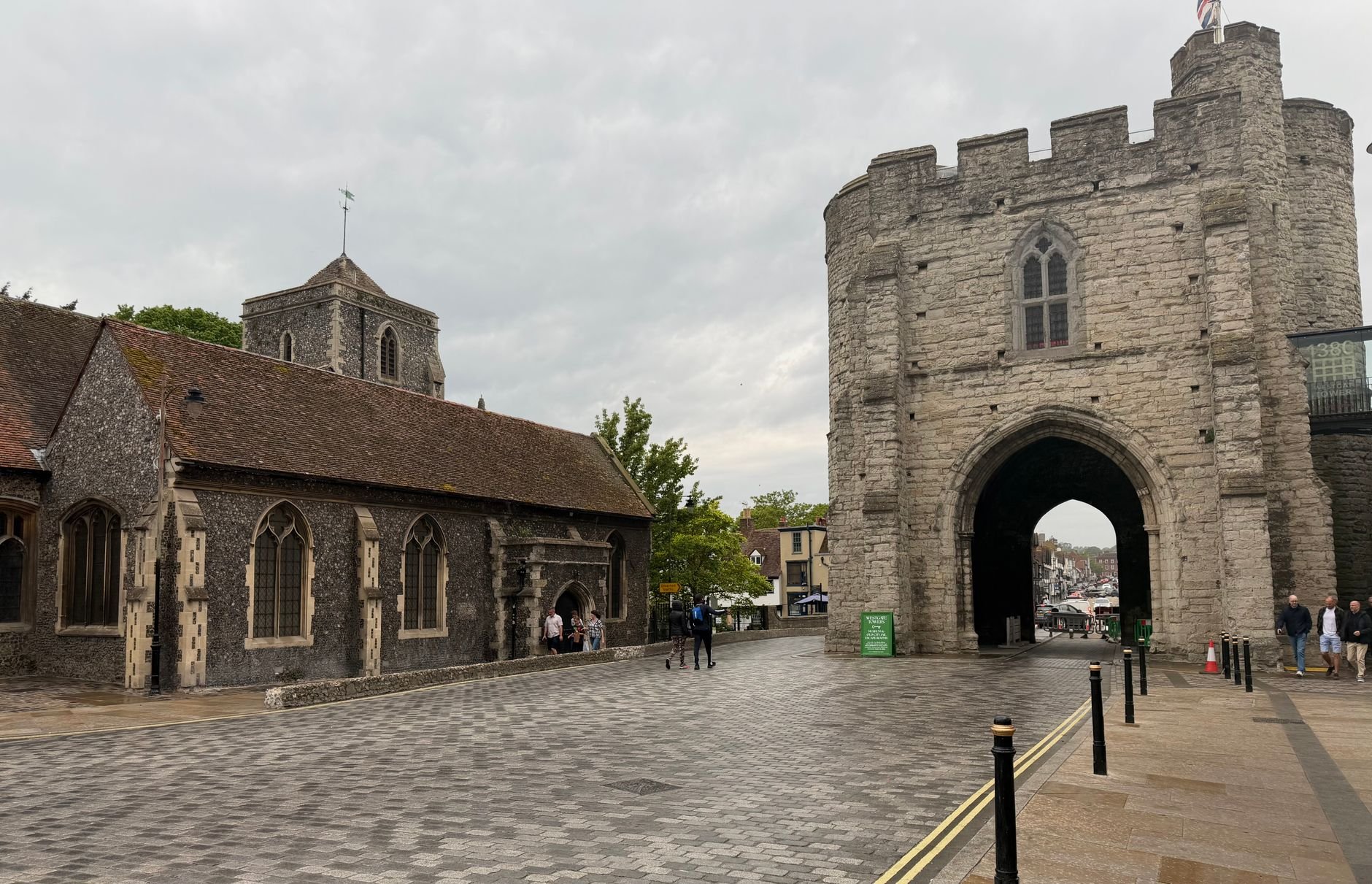 Cobbled street leading to a medieval stone gate tower with an arched passage, beside a flint-and-stone church with pointed windows, under an overcast sky