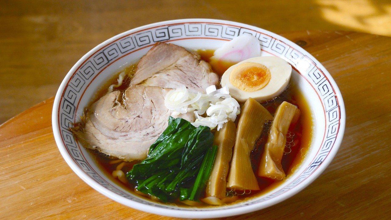 Ramen bowl with clear brown soy-coloured broth, sliced chashu pork, spinach, menma bamboo shoots, sliced white onion and a halved egg