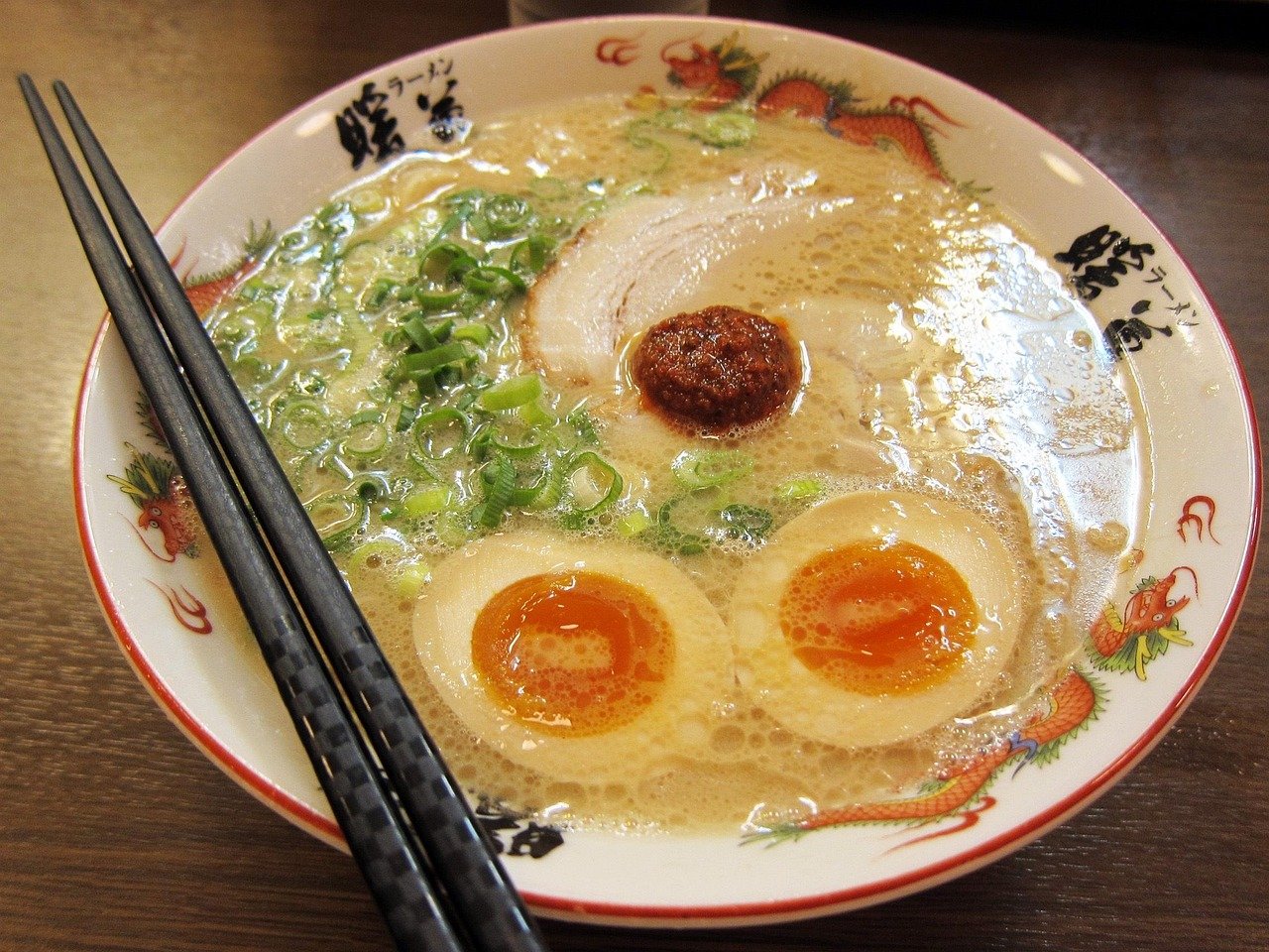 White ramen bowl with cloudy, creamy off-white broth, green onion, a slice of pork, a halved egg and a dark red paste dollop, with chopsticks resting on the bowl