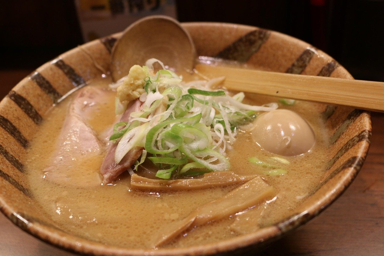 Striped brown ceramic bowl of creamy tan cloudy broth with sliced green onion, pork slices, bamboo shoots and a halved egg, with a wooden ladle and chopsticks