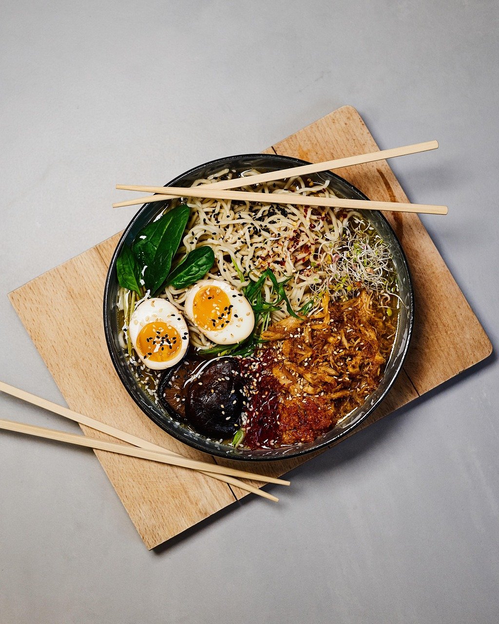 Overhead view of a ramen bowl with noodles, a halved soft-boiled egg, spinach, sprouts, mushrooms and shredded topping, set on a wooden board with two pairs of chopsticks