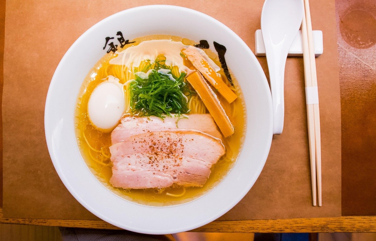 Overhead view of a white bowl with clear golden broth, sliced chashu pork, a halved egg, green onion and bamboo shoot strips, beside a spoon and chopsticks