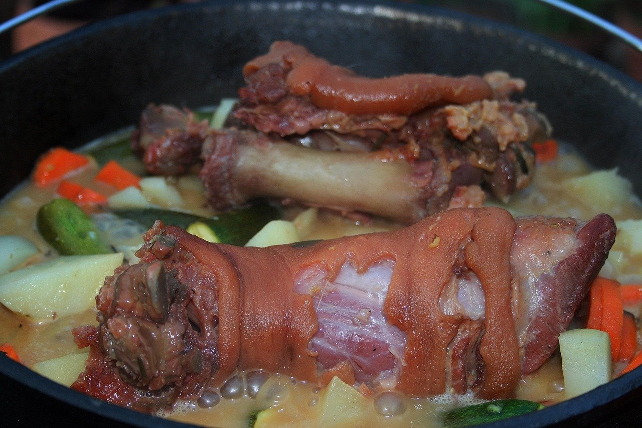 Black pot of broth with pork knuckle pieces, potatoes, carrots and cucumber pieces