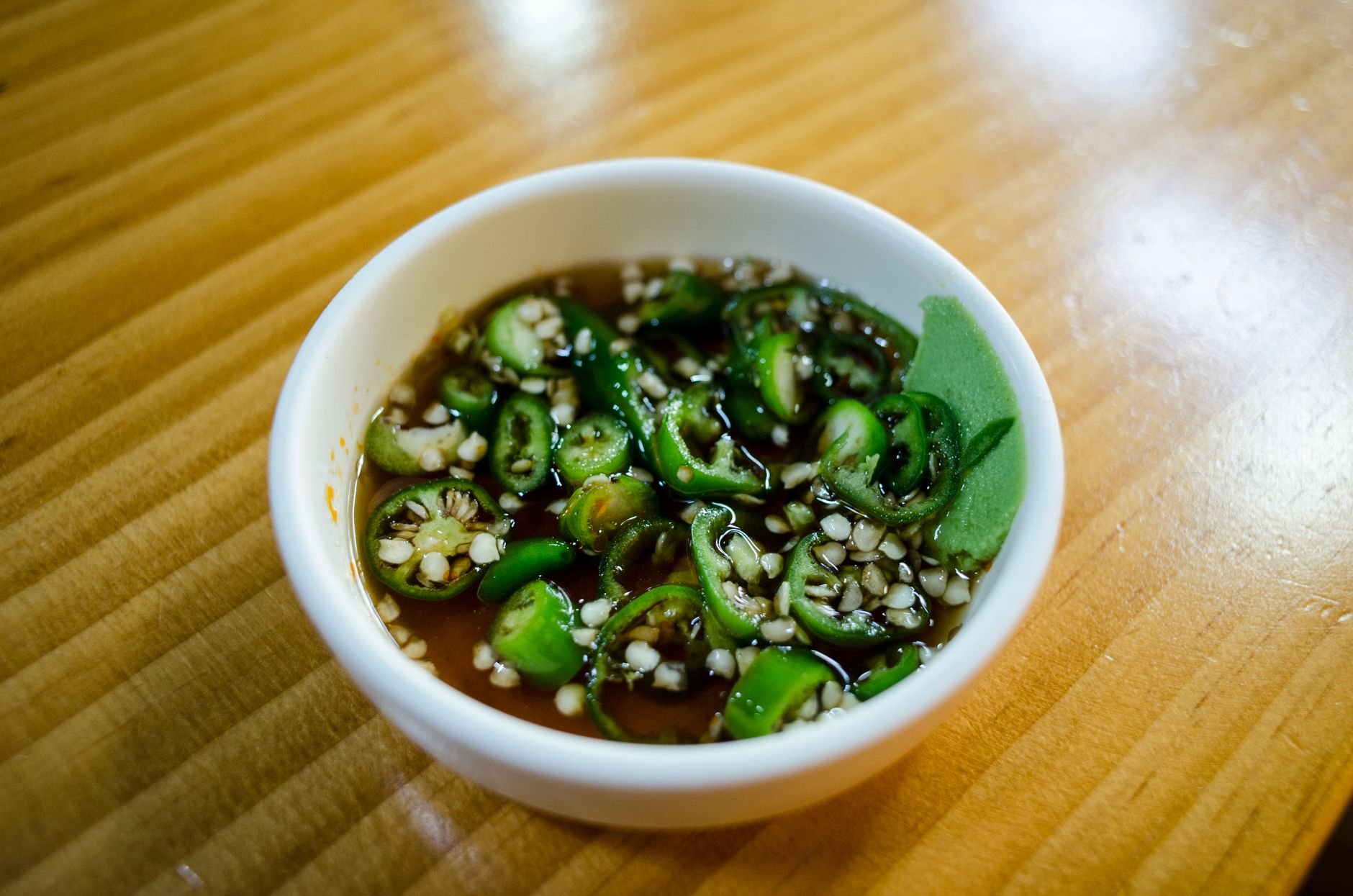 Small white bowl of sliced green chilies and sesame seeds in a brown liquid on a wooden table