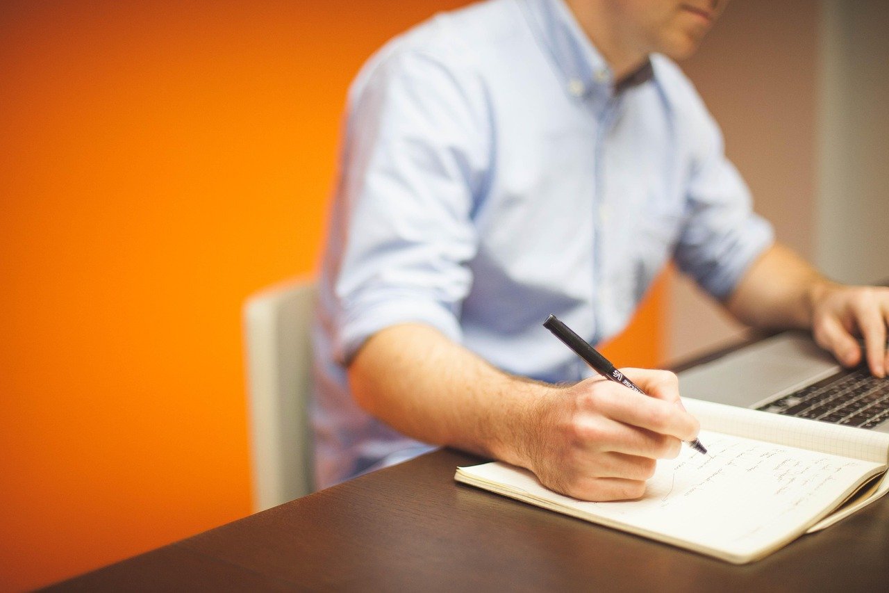 A man in a light blue shirt writing with a black pen in an open notebook, with a laptop beside him and an orange wall behind