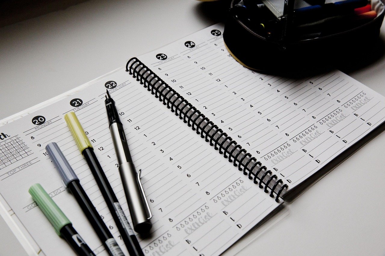 An open spiral planner with numbered hour lines and date markers, with several felt-tip pens and a pen cup on a light desk