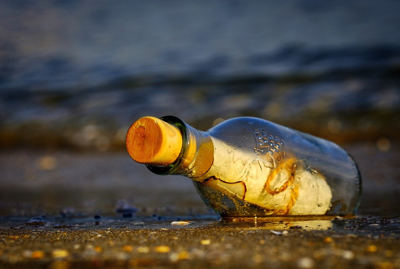 A glass bottle with a wooden-colored cork lying on wet sand at the edge of the water, with soft waves in the background