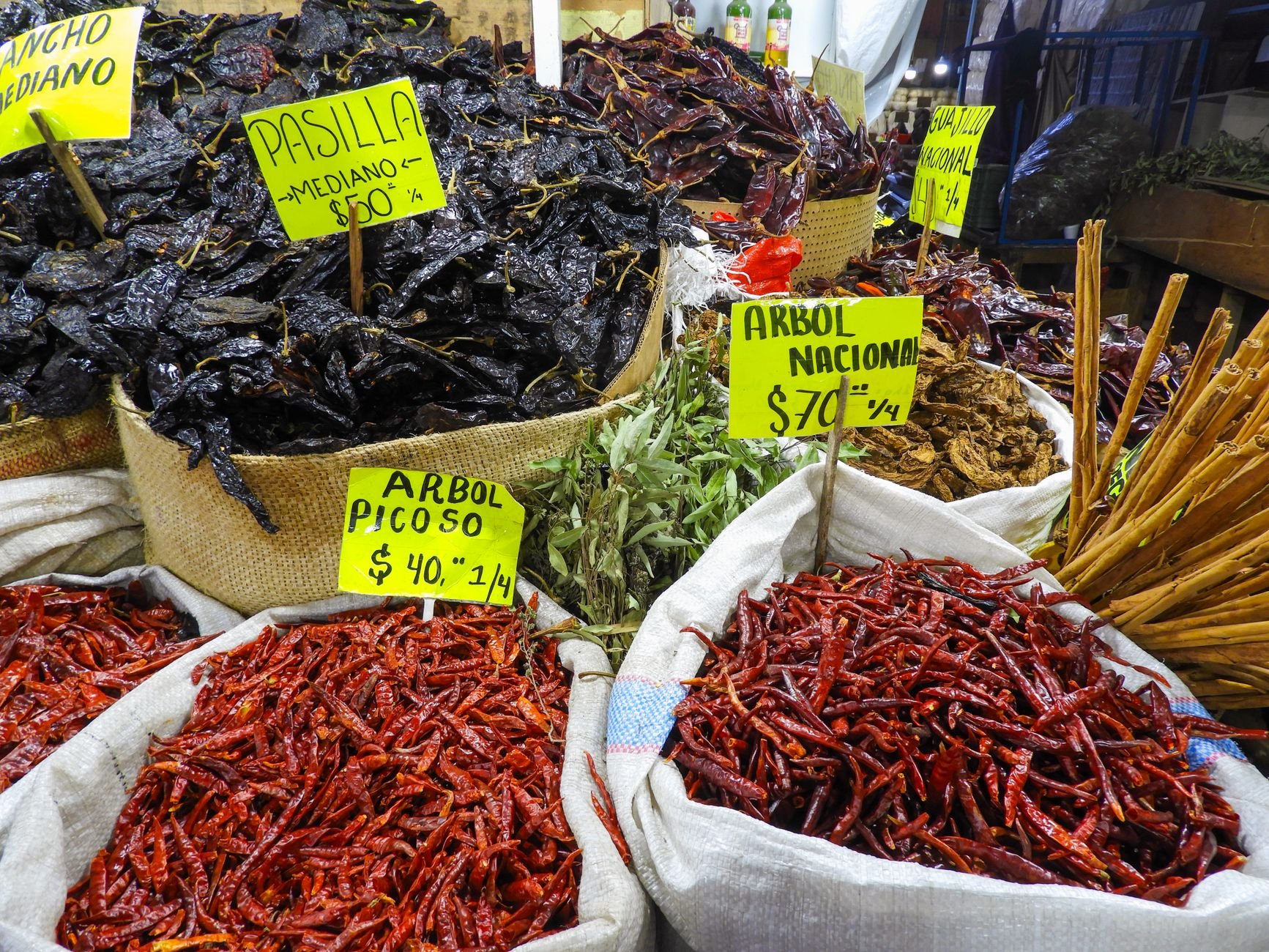 Dried red chiles and dark dried chiles in burlap and white sacks at a market stall, with handwritten yellow price signs