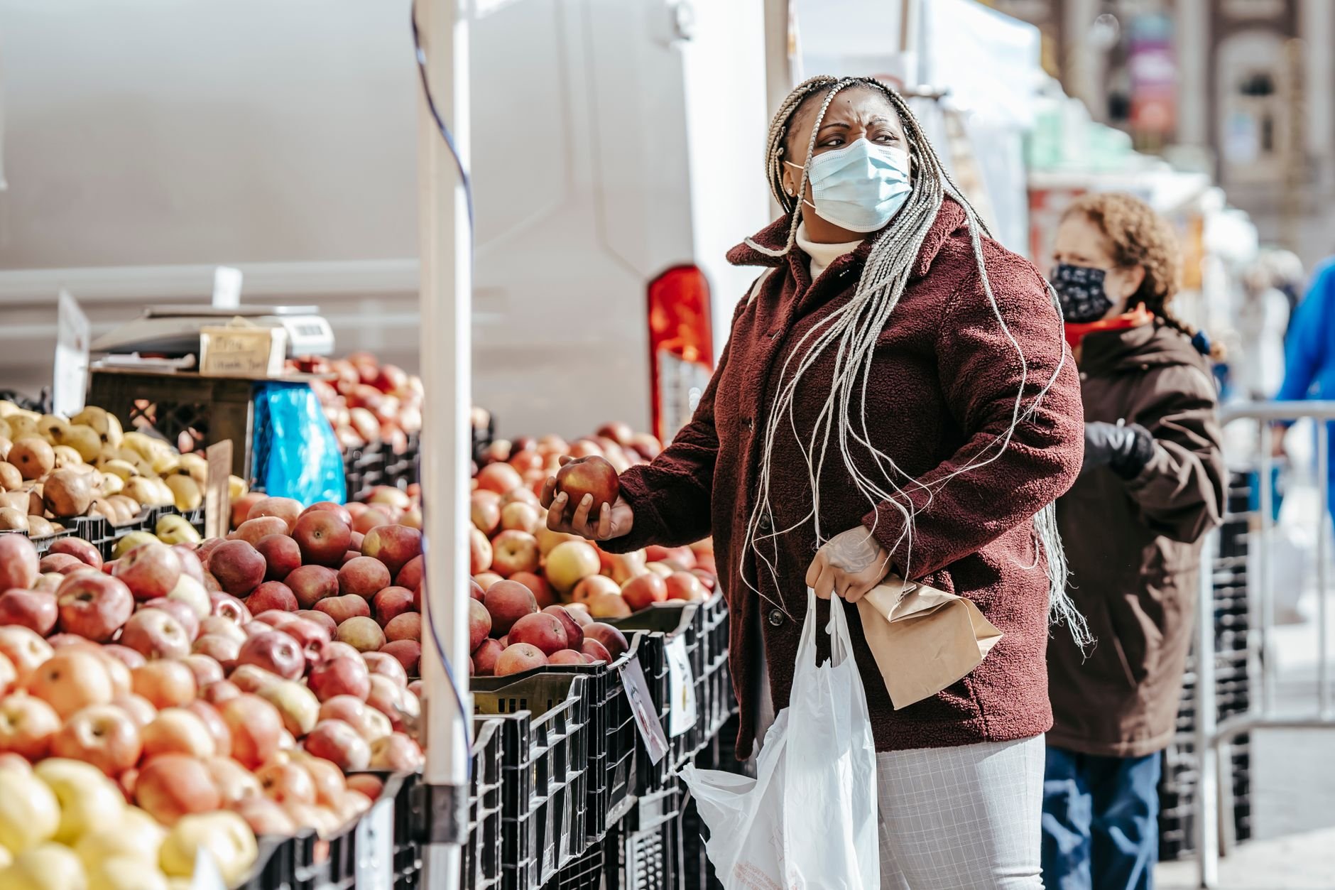 Woman in a face mask and a maroon coat choosing an apple from a crate at a market stall, with another shopper behind her