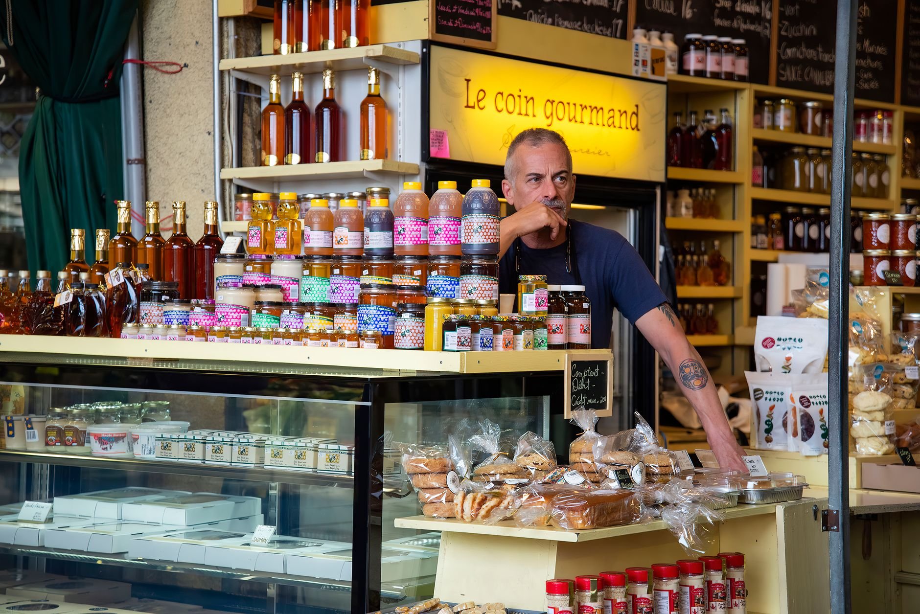 Vendor in a dark shirt behind a counter with jars of jam and preserves, bottles of oil and vinegar, and packaged bread