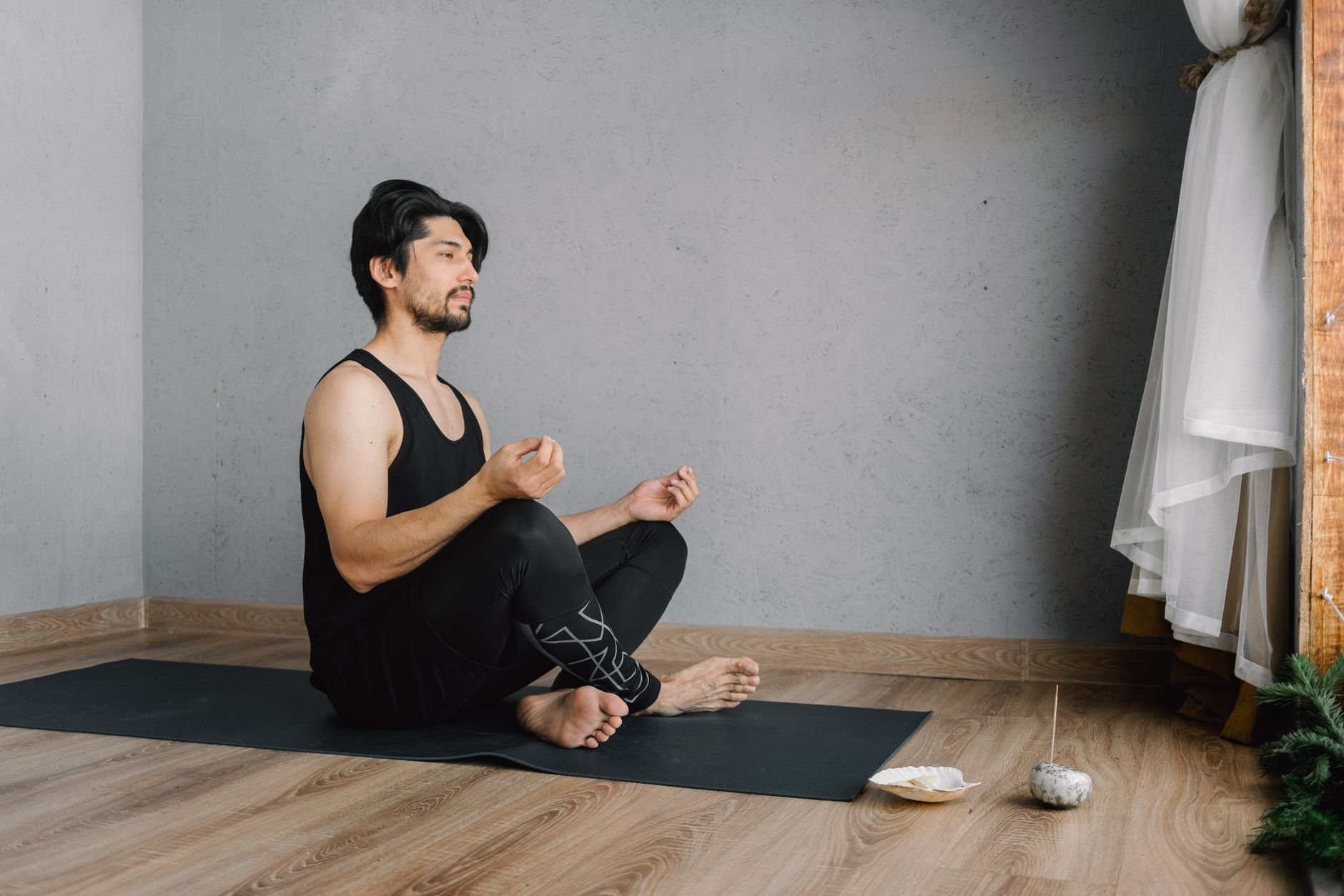 Man with short dark hair and a beard, eyes closed, in a black sleeveless top and black pants, seated cross-legged on a black mat on a wooden floor with hands held in front of his body near his knees, grey wall behind him