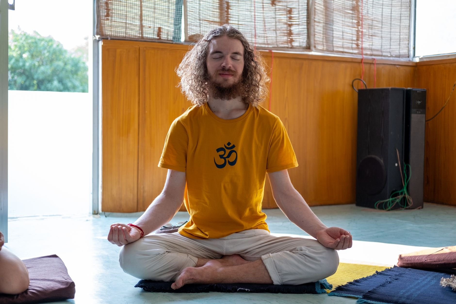 Man with long curly hair and a beard, eyes closed, wearing a yellow t-shirt with a black Om symbol, seated cross-legged on a dark mat with hands resting near his knees, yellow wood-paneled wall and a black speaker behind him