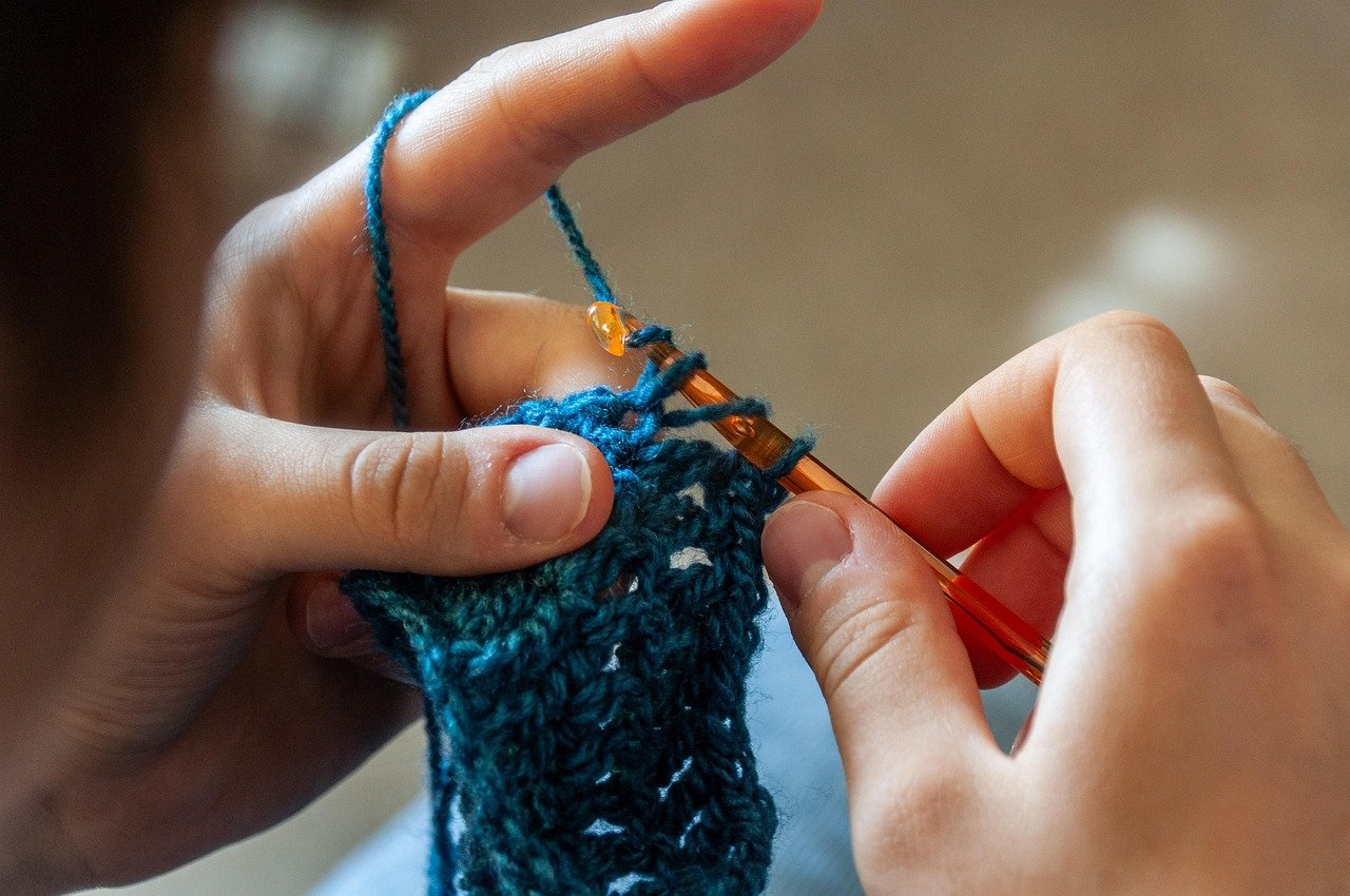 Hands crocheting a dark teal piece with an orange crochet hook