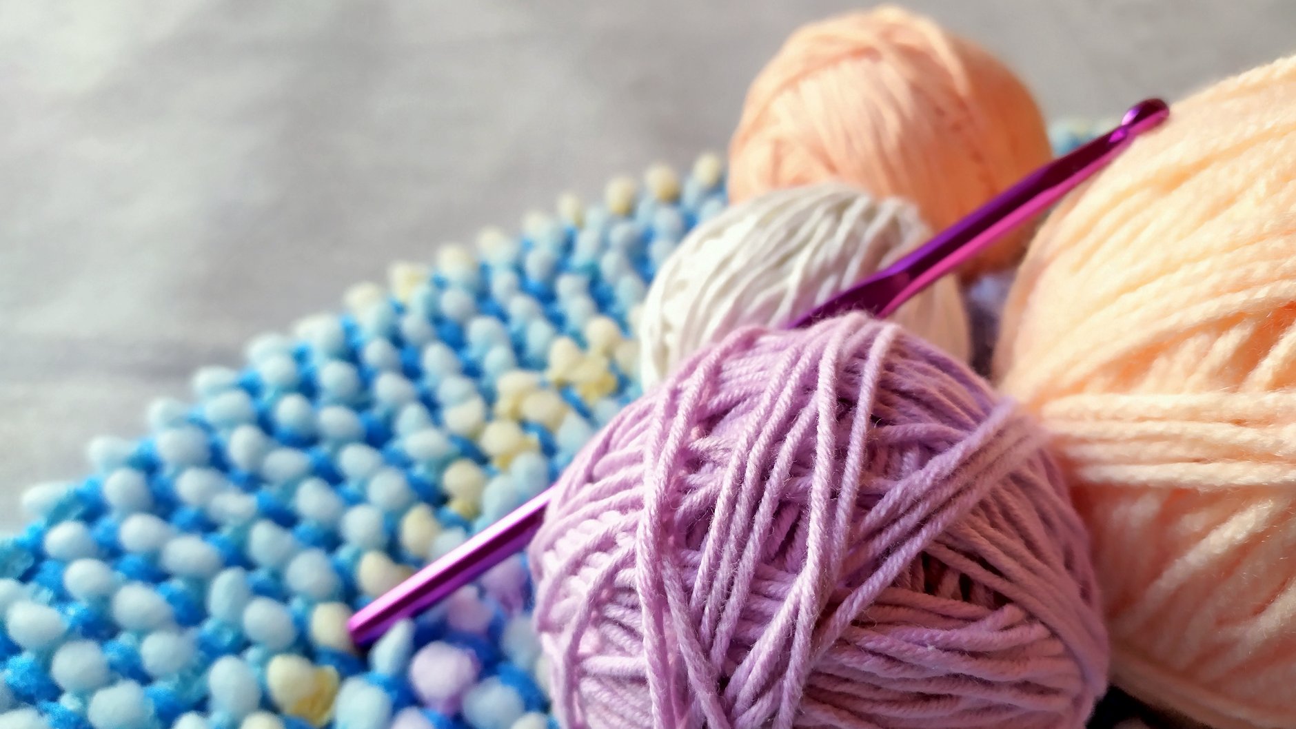 Lilac, peach, and white yarn balls with a purple crochet hook resting on a textured blue crocheted fabric