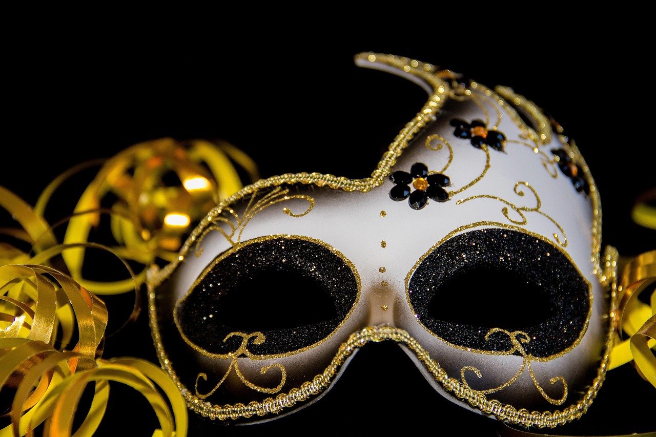 White Venetian half mask with gold trim, black glitter eye openings, and black floral detailing against a dark background