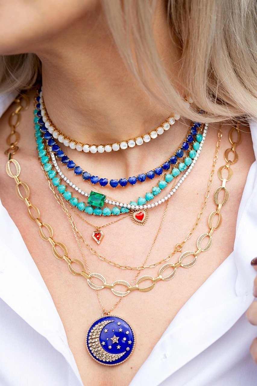 Several layered gold and beaded necklaces, including a blue enamel moon pendant, worn over a white collared shirt