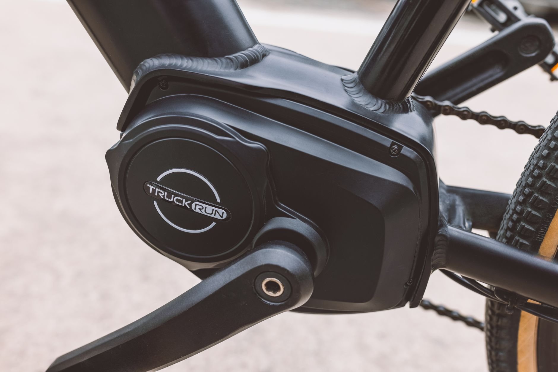Black mid-drive motor unit at the bottom bracket of a black e-bike frame, with a chainring and tyre visible
