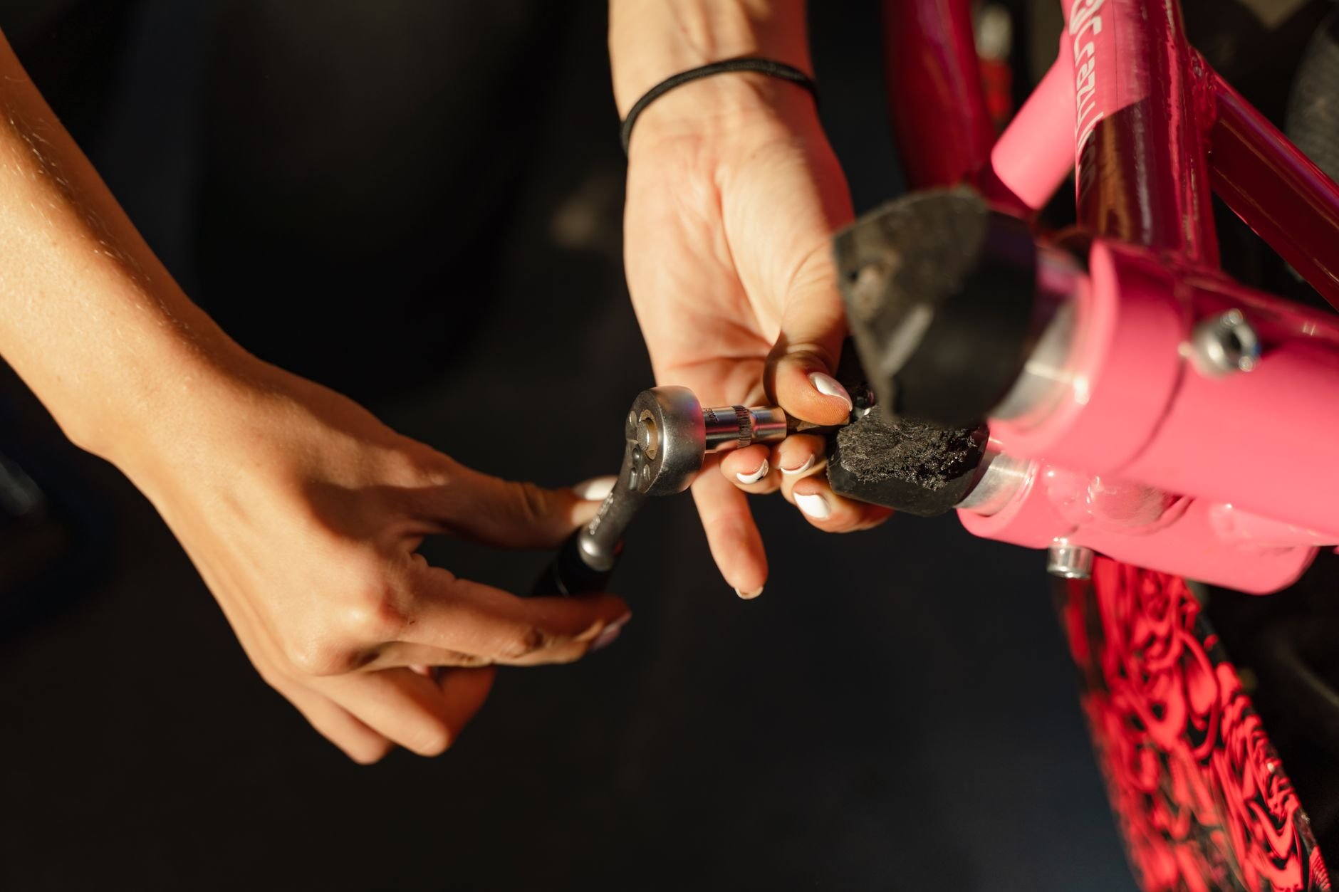 Hands holding a small metal tool at the end of a pink bicycle fork, with a dark background