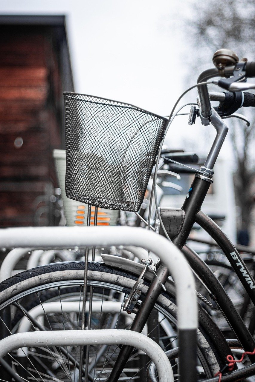 Black city bike with a wire basket and bell parked in a metal rack beside other bikes