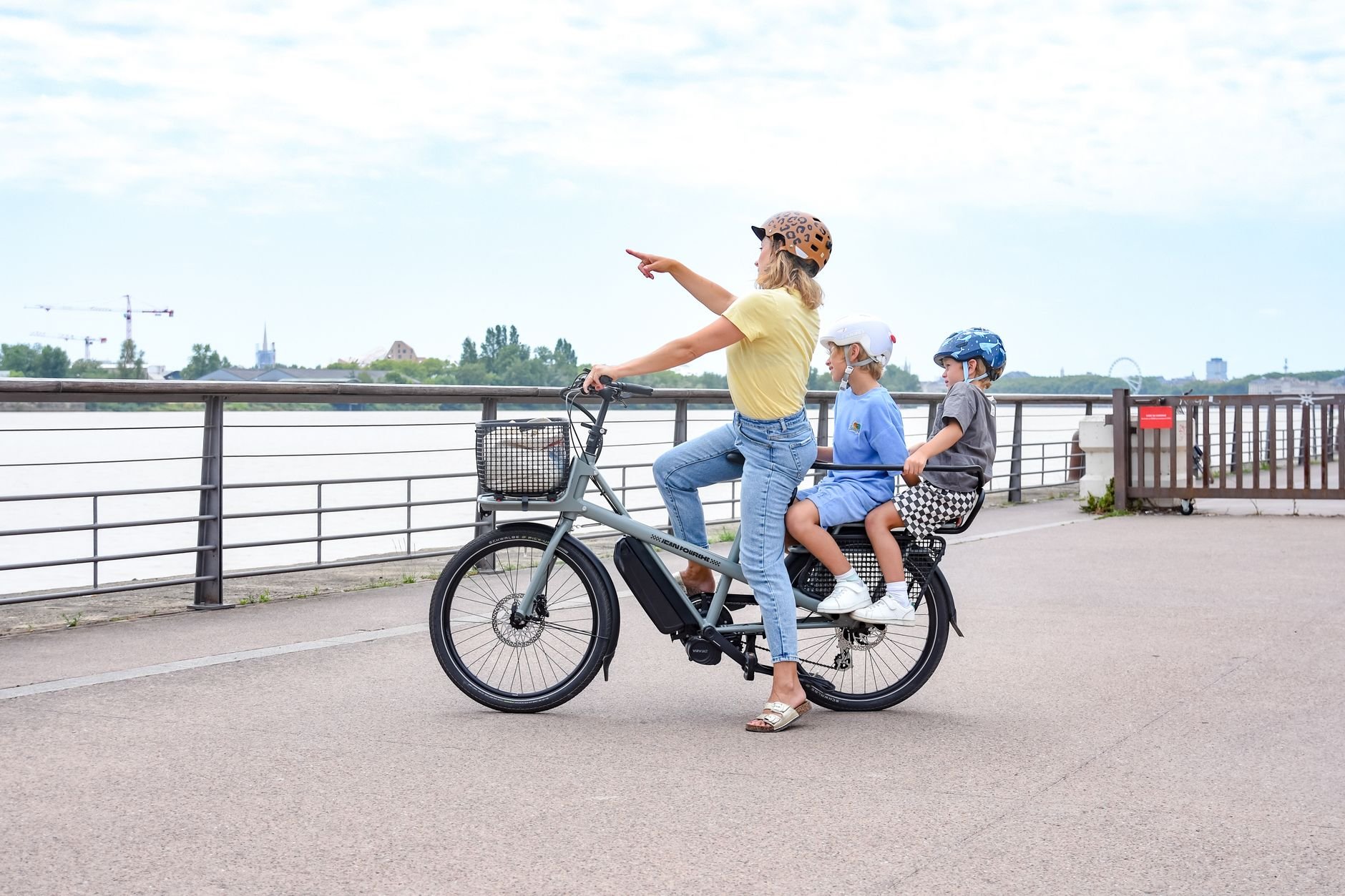 Woman in a yellow top and two children riding a gray electric bike with a front basket along a riverside promenade