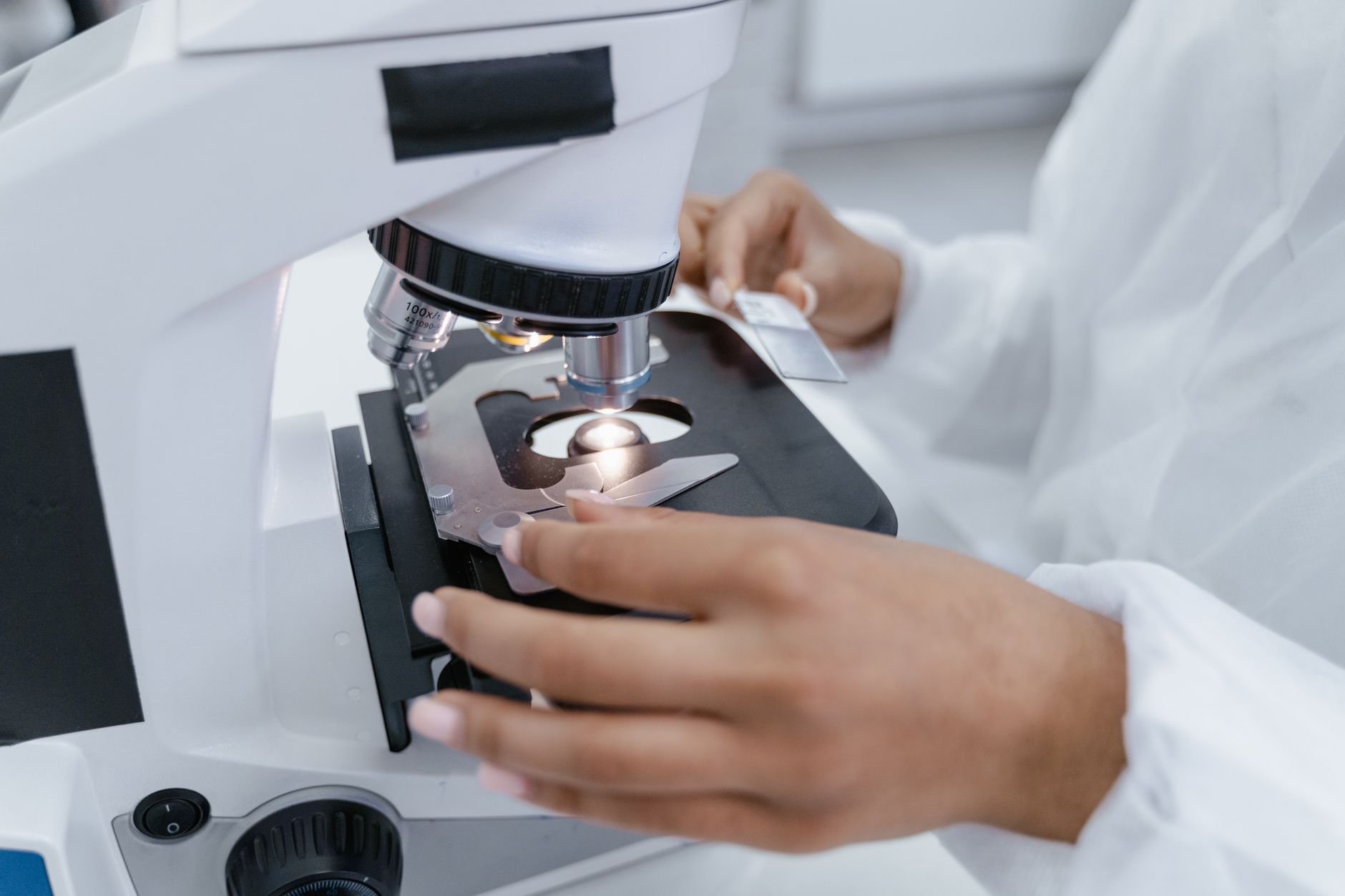 A laboratory microscope viewed from the side, with a glass slide on the stage and a light shining through it, while a person in a white lab coat adjusts the slide with one hand and holds another slide in the other.