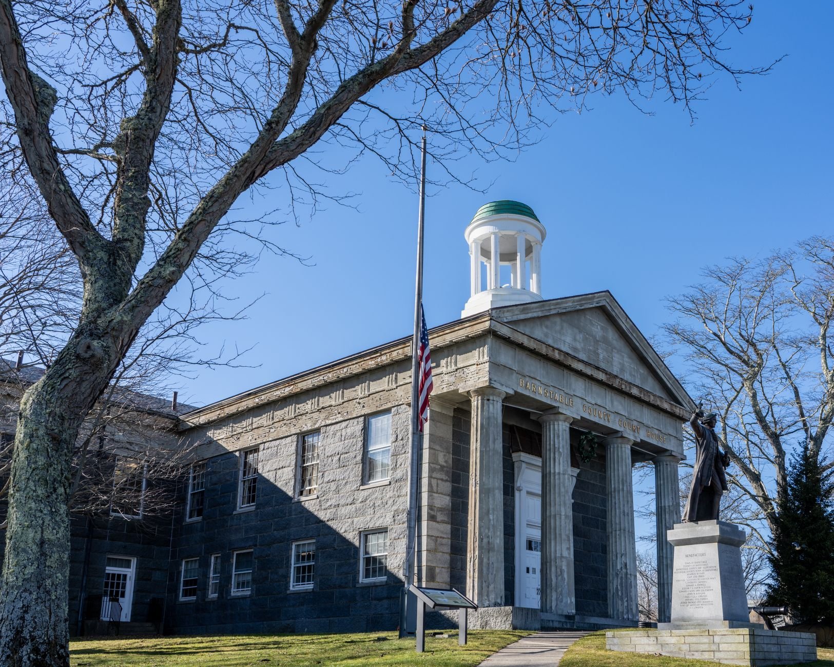Flag hanging on a pole at half-staff in front of a stone building with a domed cupola and a statue on the right