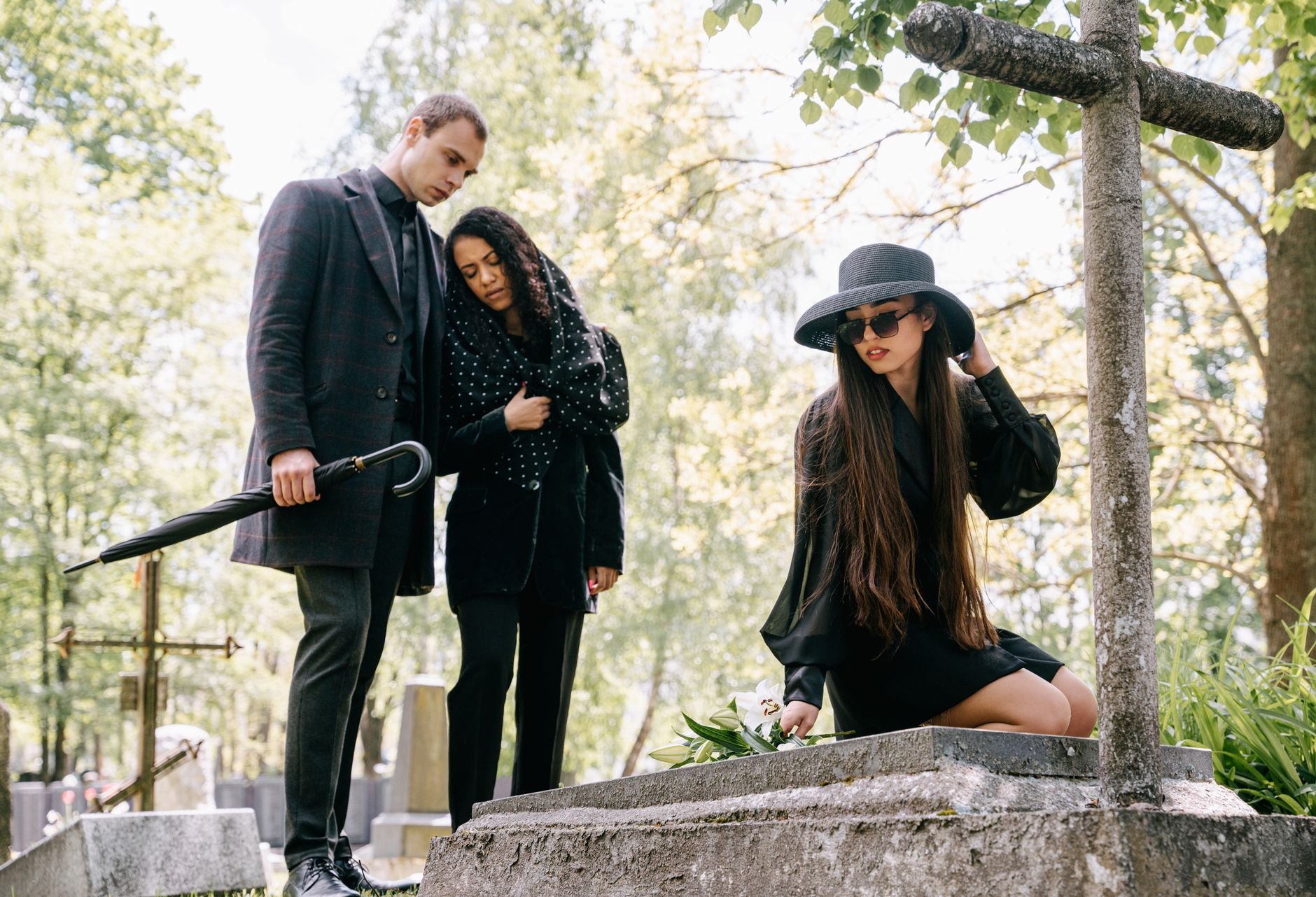 A woman in a black hat kneels at a grave and lays flowers on a stone, while a man holding an umbrella and a woman in black stand nearby among trees