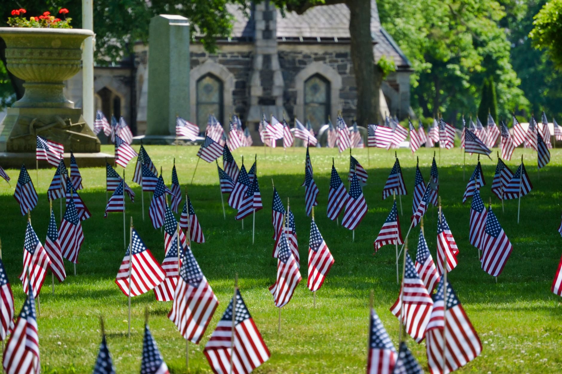 Many small American flags on sticks planted across a green cemetery lawn, with a stone building and trees in the background