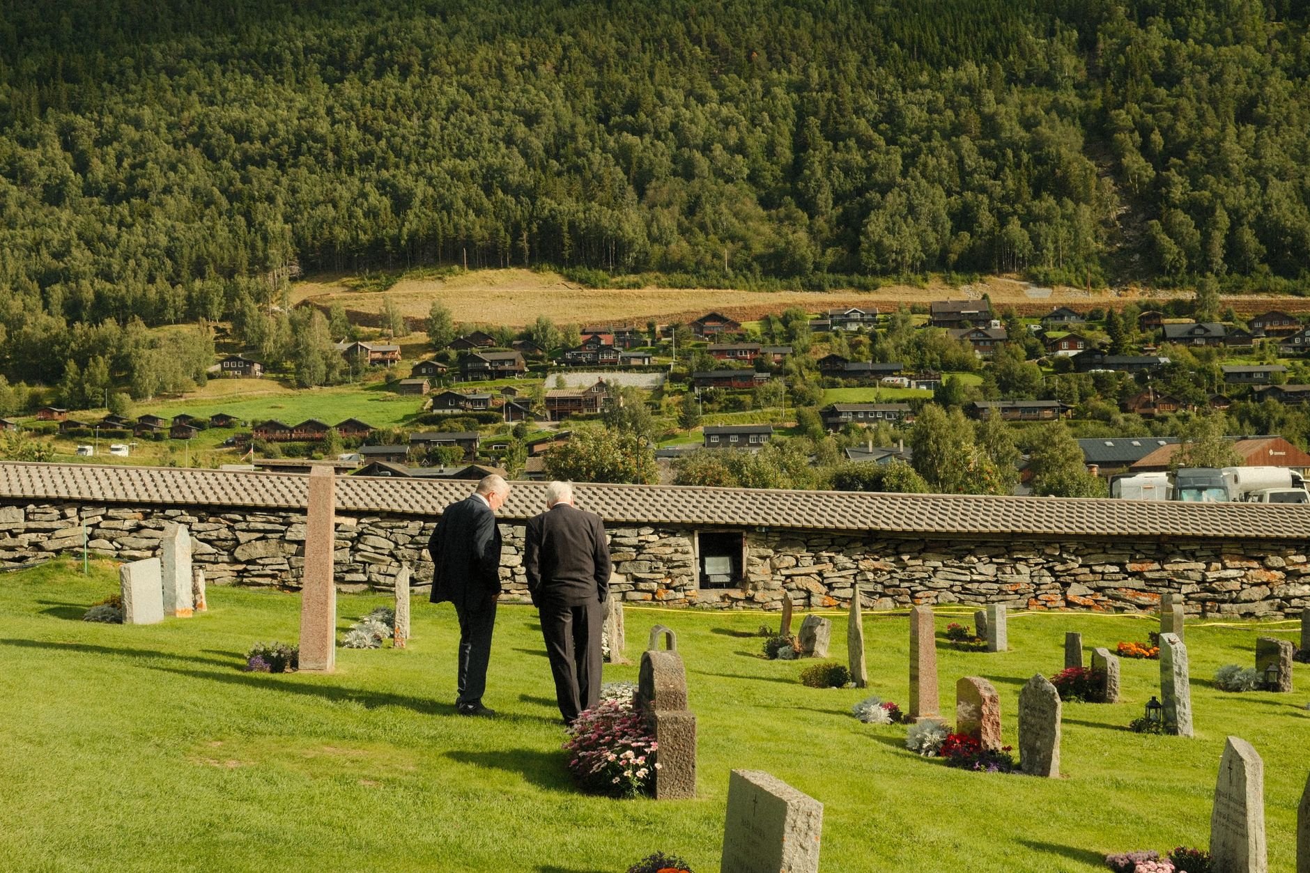 Two men in dark coats walk away across a grassy cemetery among flower-decorated grave markers, with a stone wall and a forested valley behind