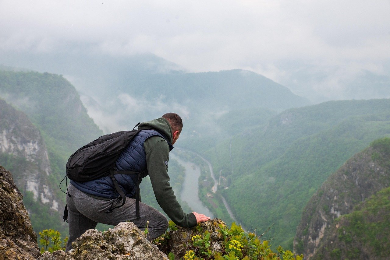 A hiker in a dark backpack kneels on a rocky outcrop above a misty river valley with forested green slopes.