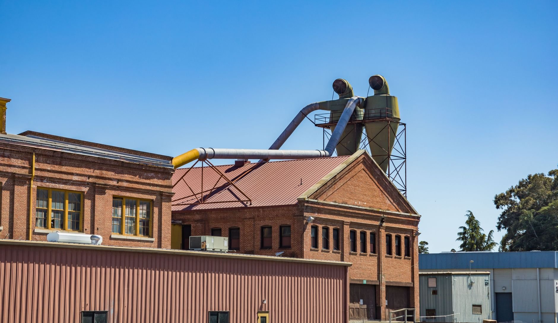 Two cylindrical metal units on a steel stand above a brick building, connected by pipes and ducting under a clear blue sky