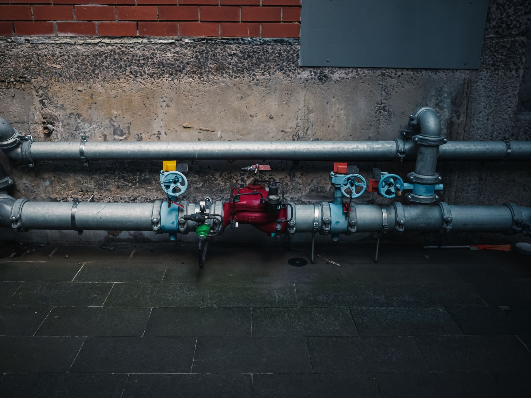 Galvanized steel pipes with red and blue valves and a red pump fixed along a grey wall above a dark paved floor