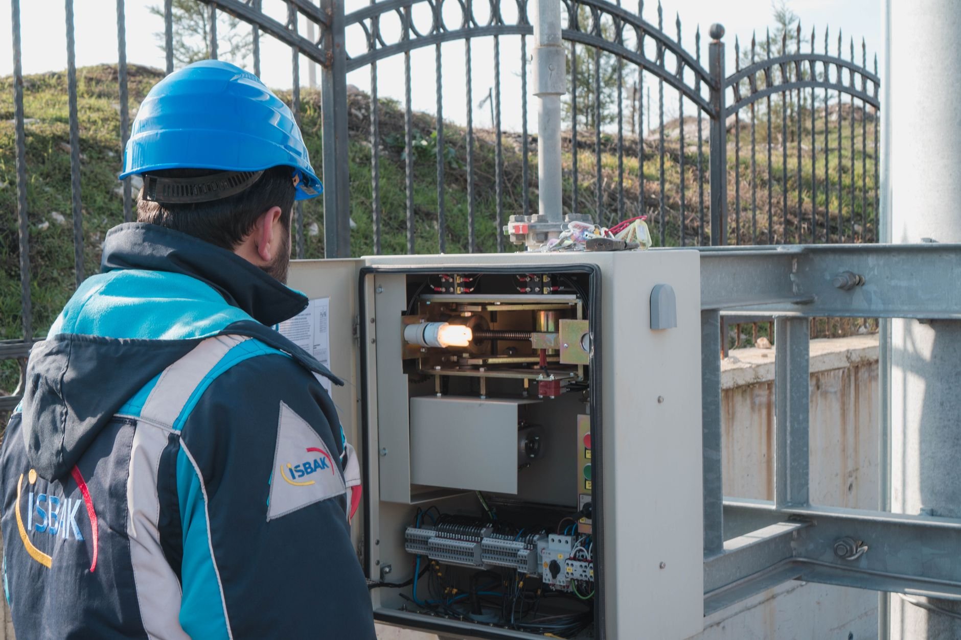 Technician in a blue hard hat and jacket looking into an open equipment cabinet beside a metal gate