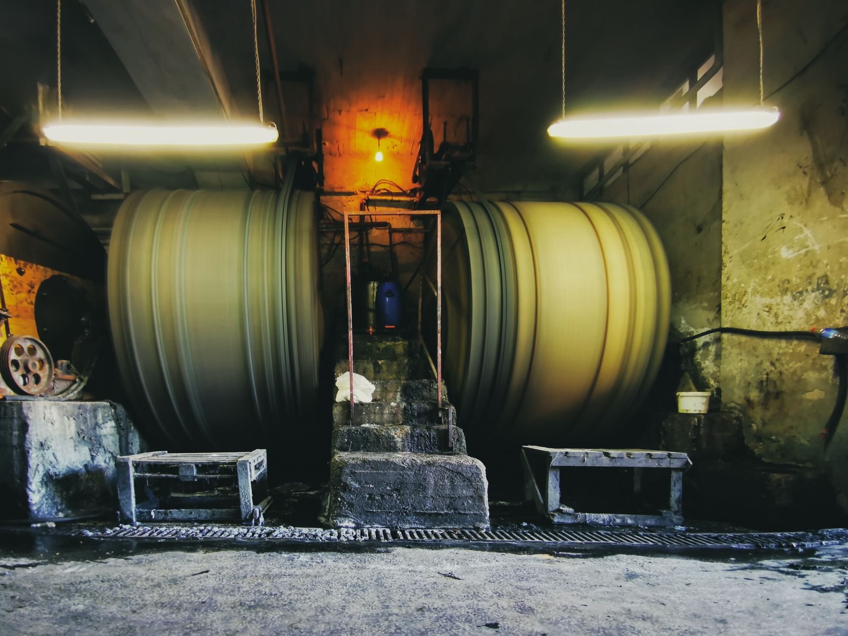 Two large cylindrical drums, one shown with motion blur, in a dim industrial room with overhead lights and a small workbench in front