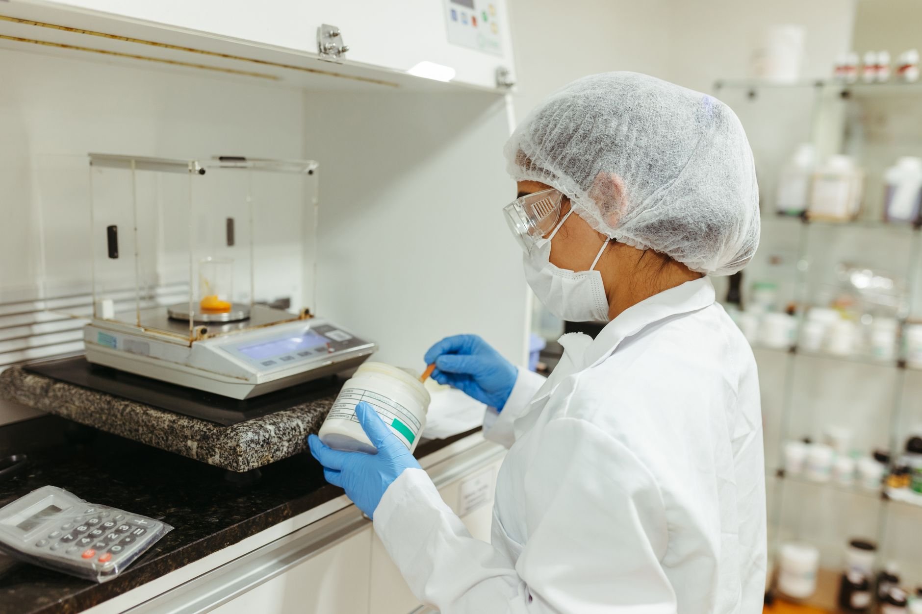 Lab worker in a white coat, hair net, mask and blue gloves weighing a white jar of powder on a laboratory balance