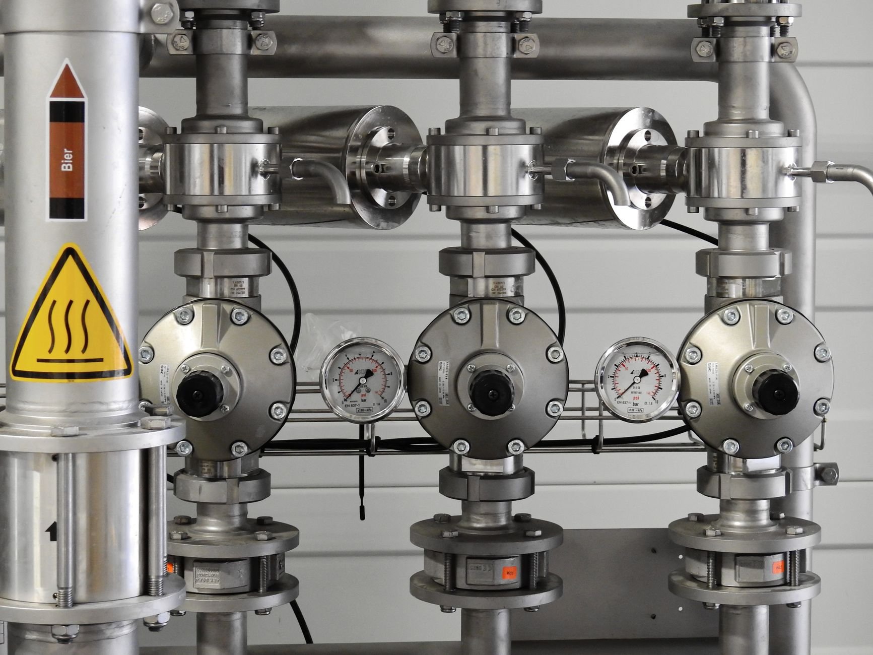 Stainless steel pipe manifold with three valves, two pressure gauges, and a yellow hot-surface warning sign on a vertical pipe