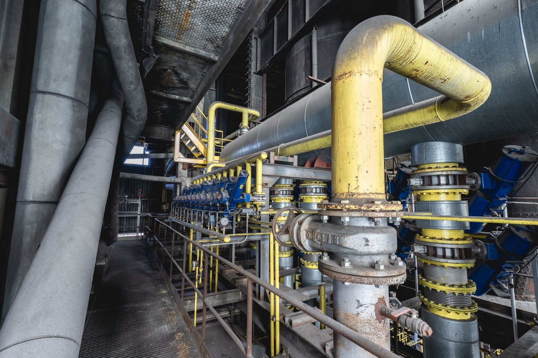 Large grey pipelines with a yellow pipe bend, flanged connections, blue valves, and a metal walkway with railings inside an industrial building