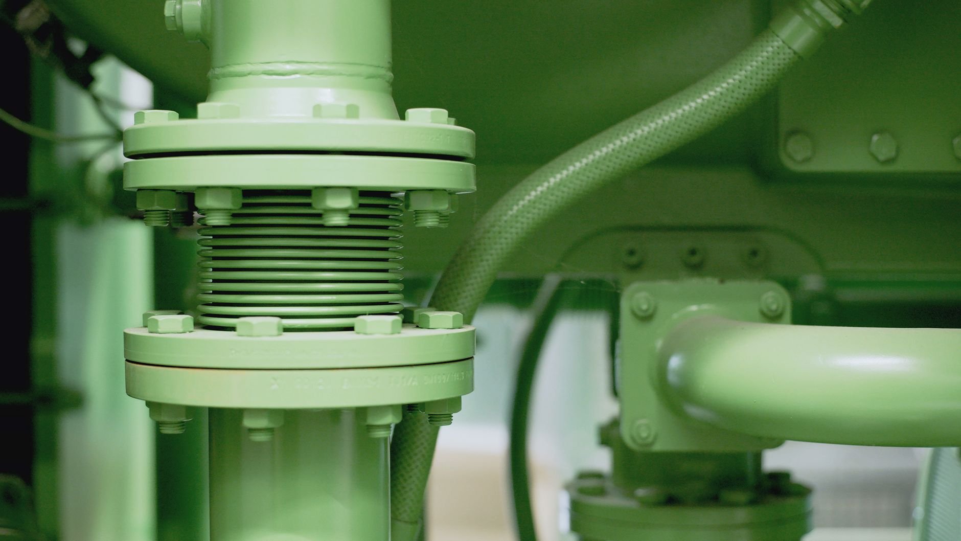 Close-up of a green flanged pipe connection with a metal expansion bellows section and a green flexible hose running alongside