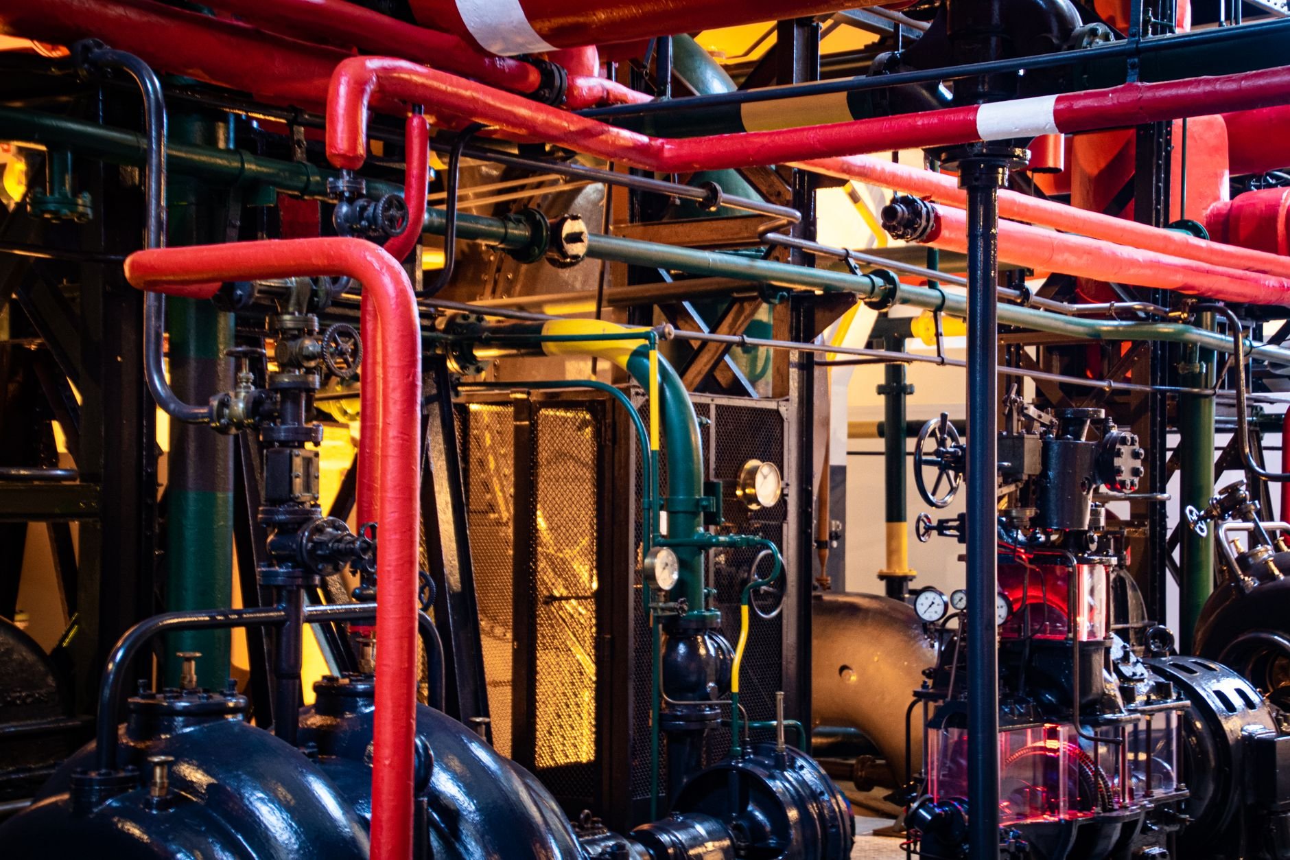 Dense industrial pipework with red and grey pipes, pressure gauges, valves, and dark machinery housings in a machine room